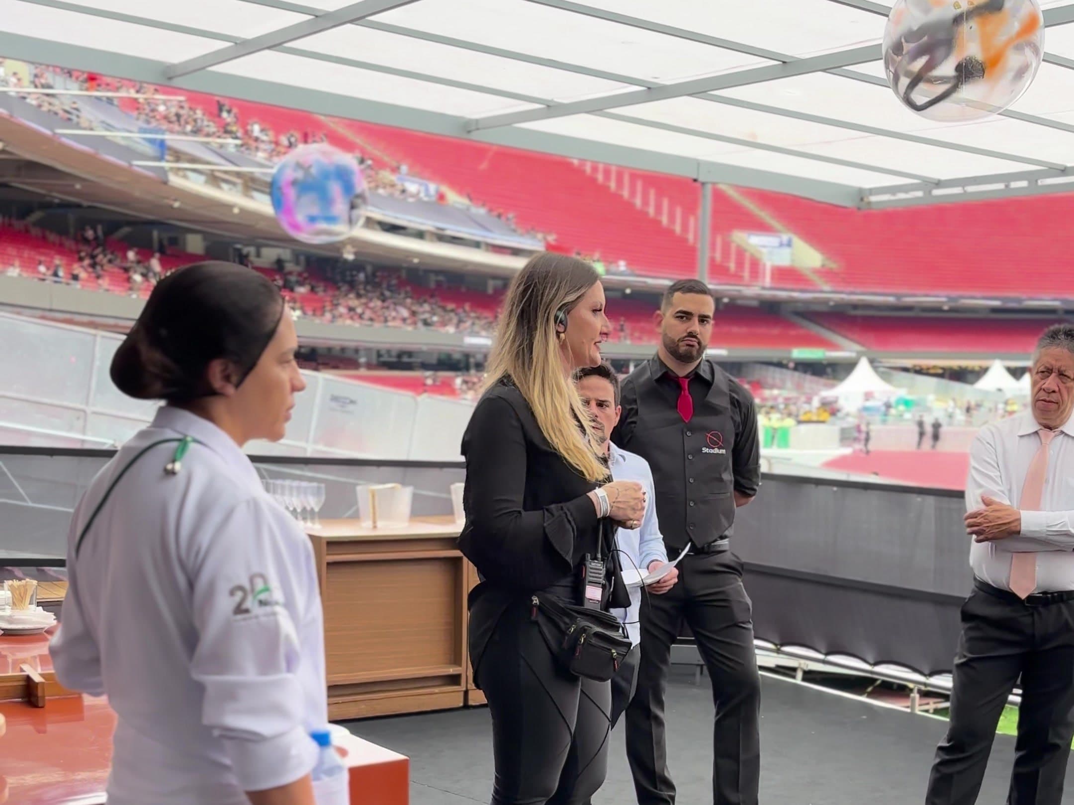 Coordenadora com rádio conduzindo o briefing da equipe de garçons, chefe de cozinha e maître dentro de um camarote coberto, com a arquibancada vermelha do estádio ao fundo.