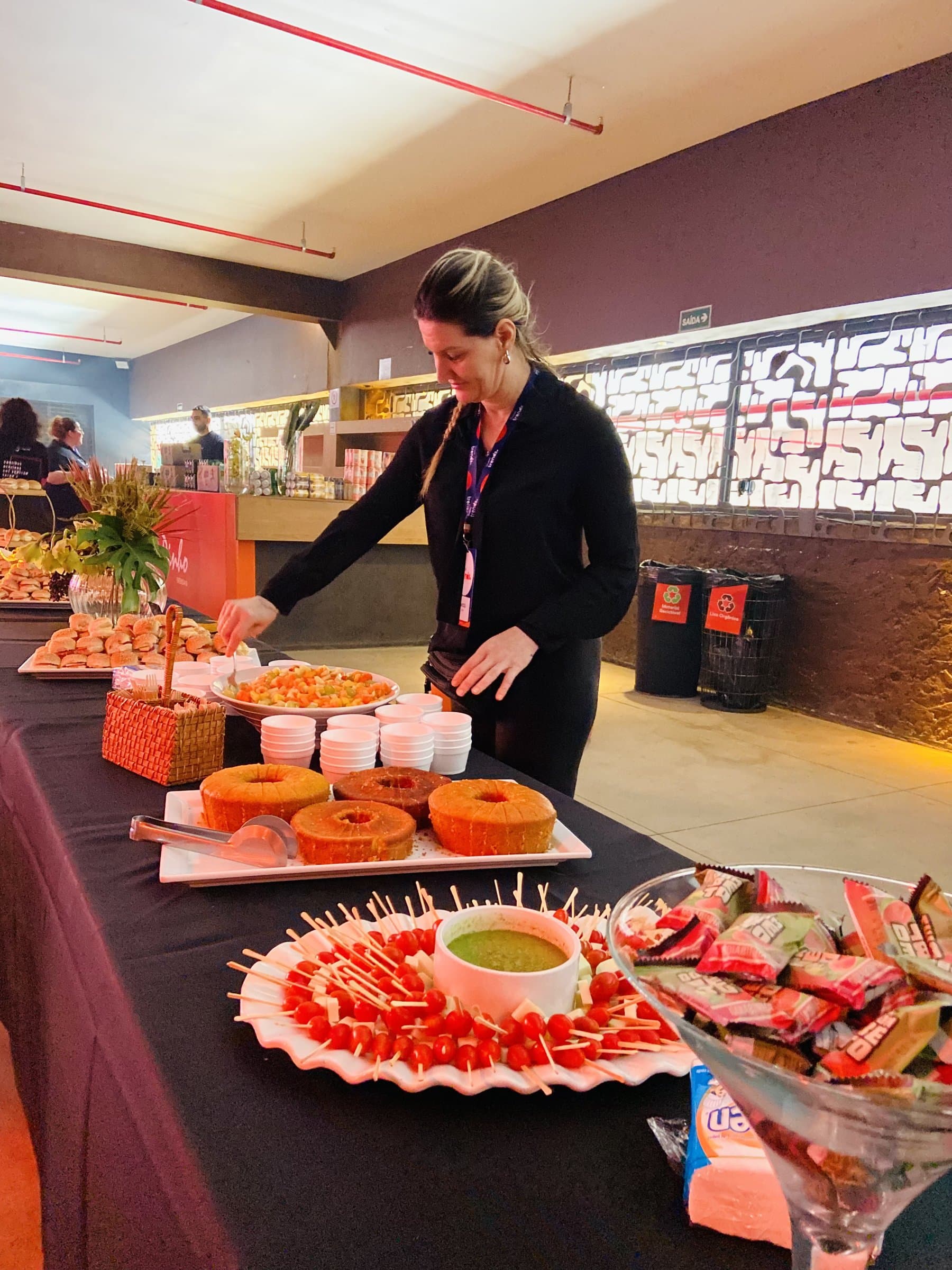 Integrante da equipe de crachá ajustando a mesa de buffet do evento, com bolos, espetinhos de tomate e salgados dispostos sobre toalha preta.