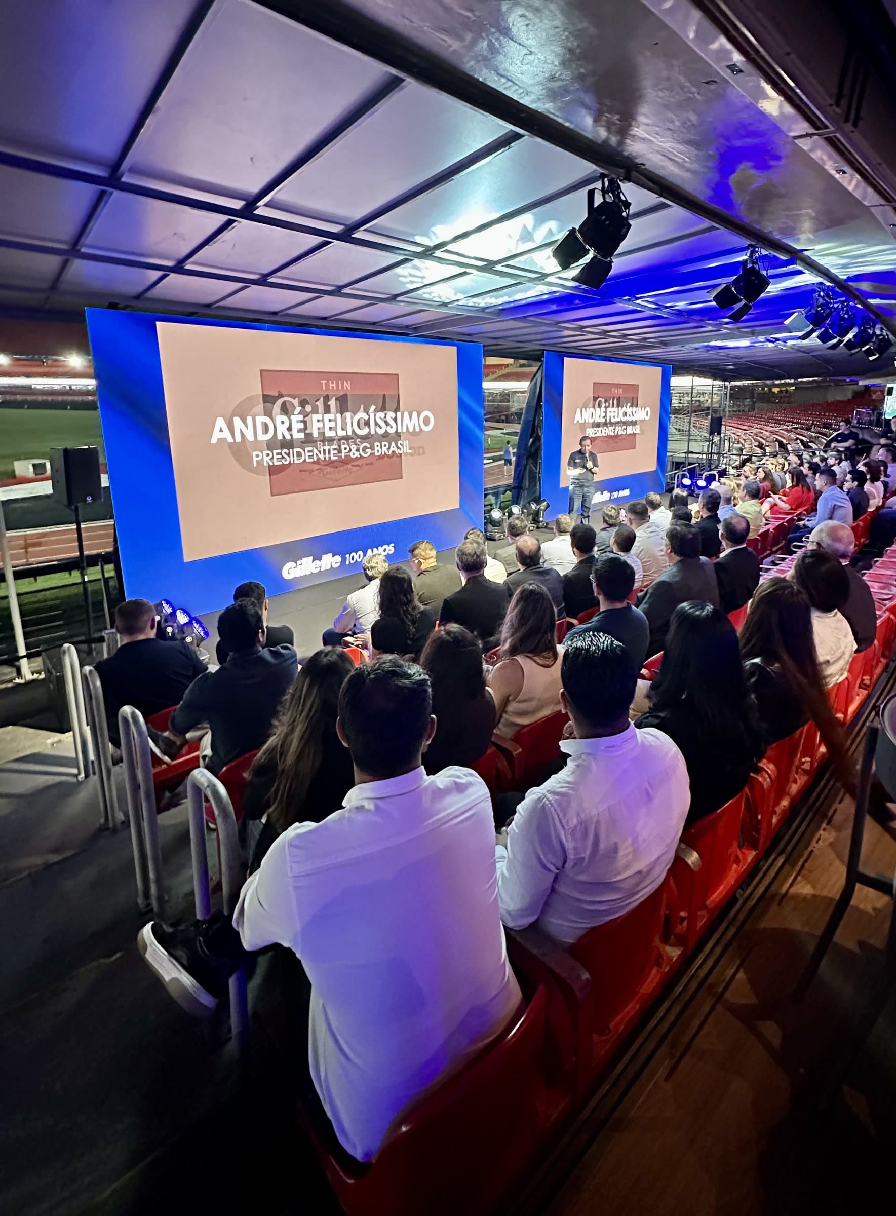Plenária corporativa na arquibancada coberta de um estádio, com o público nas cadeiras vermelhas assistindo à apresentação em dois telões de LED e o gramado ao fundo.