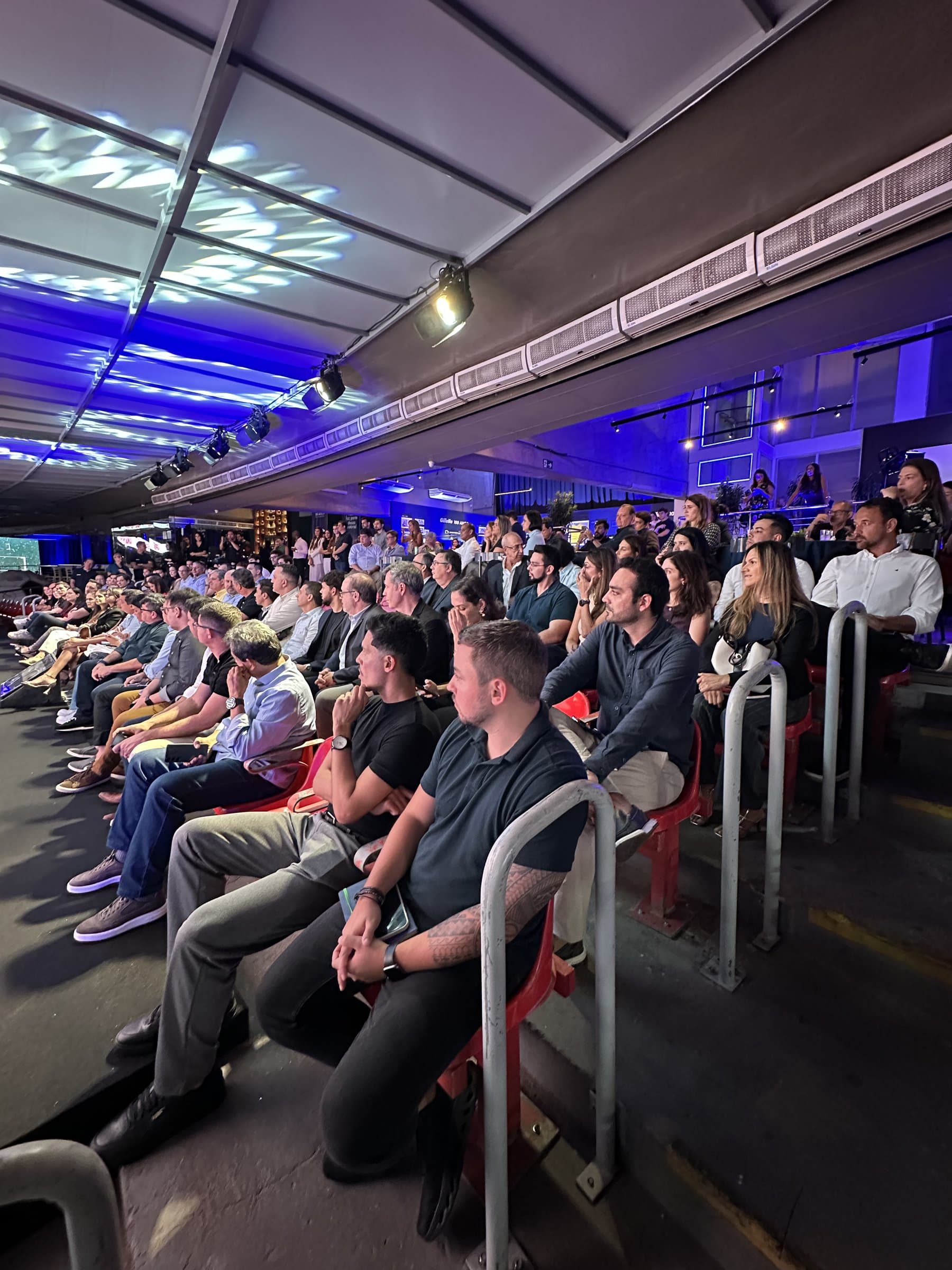 Plateia de evento corporativo sentada nas cadeiras vermelhas da arquibancada coberta de um estádio, sob iluminação cênica azul.
